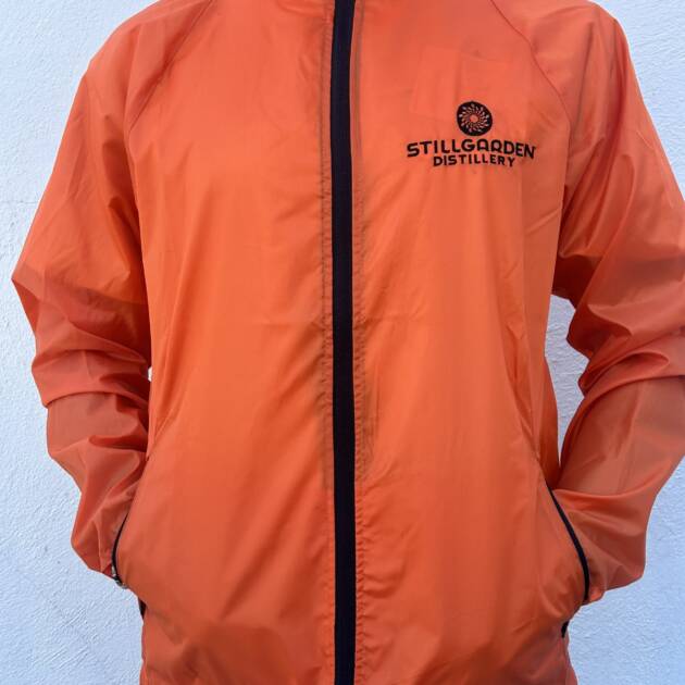 A person wears the Lightweight Jacket with Logo—an orange zip-up featuring "Stillgarden Distillery" and its logo on the left chest—while standing against a plain white background.