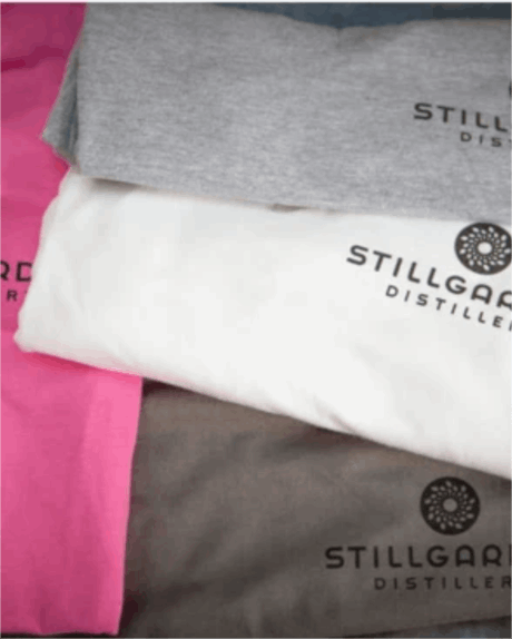Four folded Ladies short sleeve v-neck t-shirts in pink, gray, white, and brown, each featuring the "STILLGARD DISTILLERY" logo.